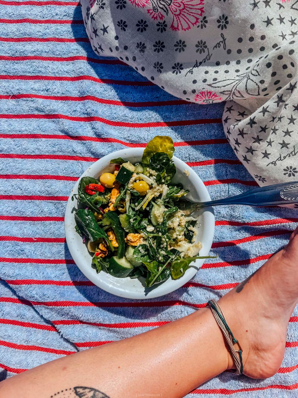 A fresh green salad in a white bowl on a striped beach towel, enjoyed outdoors on a sunny day.