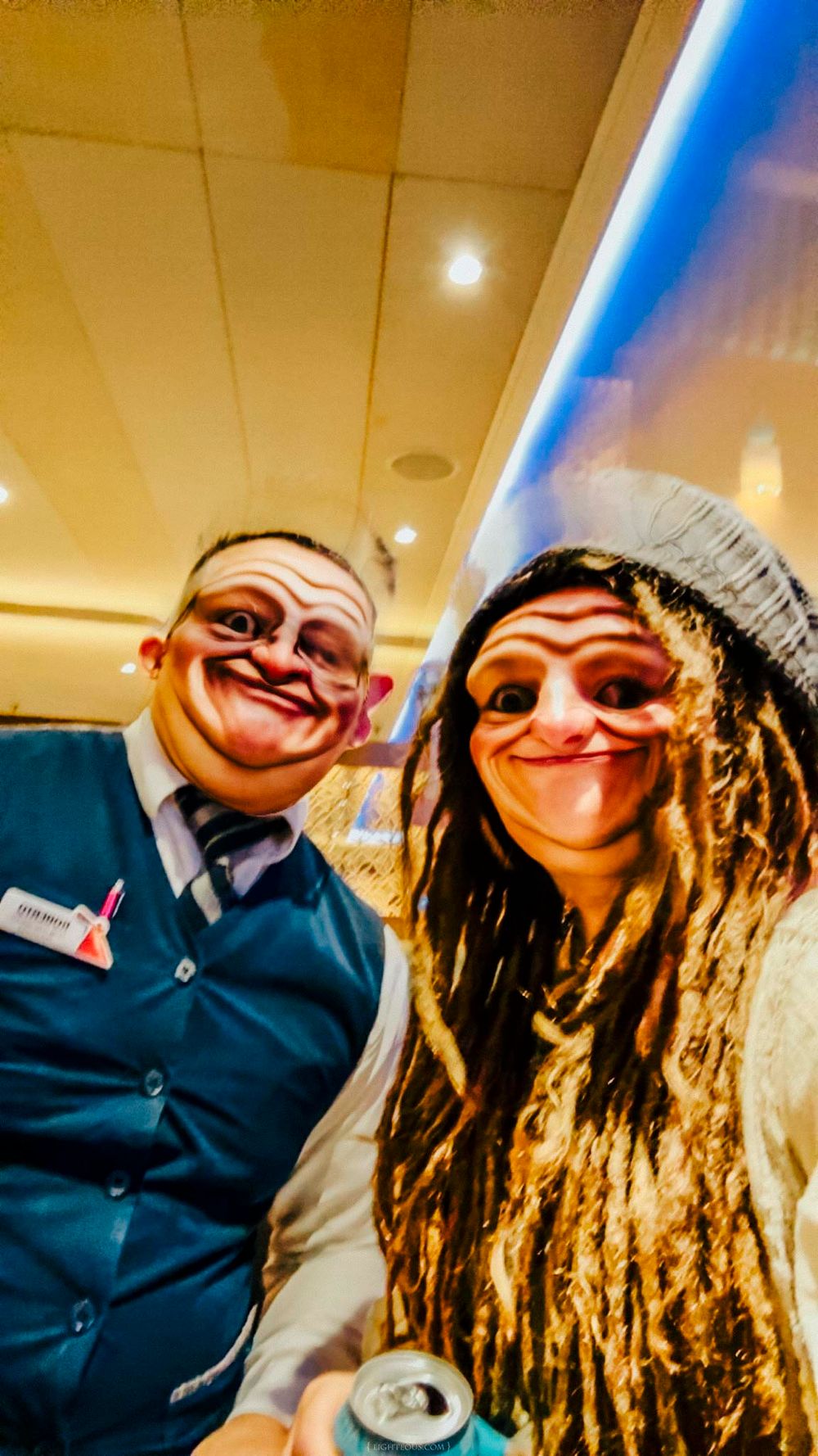 Two people wearing fun face filter masks pose for a selfie at an indoor event, one in a blue vest with a name tag.