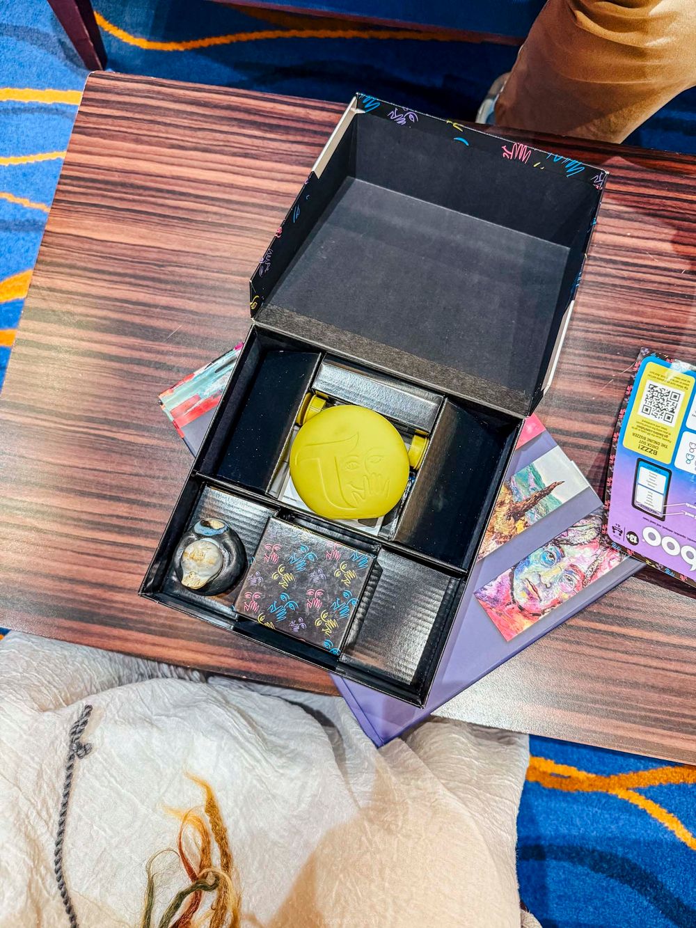 Open gift box with yellow toy, floral cards, and colorful accessories on wooden table with blue carpet.