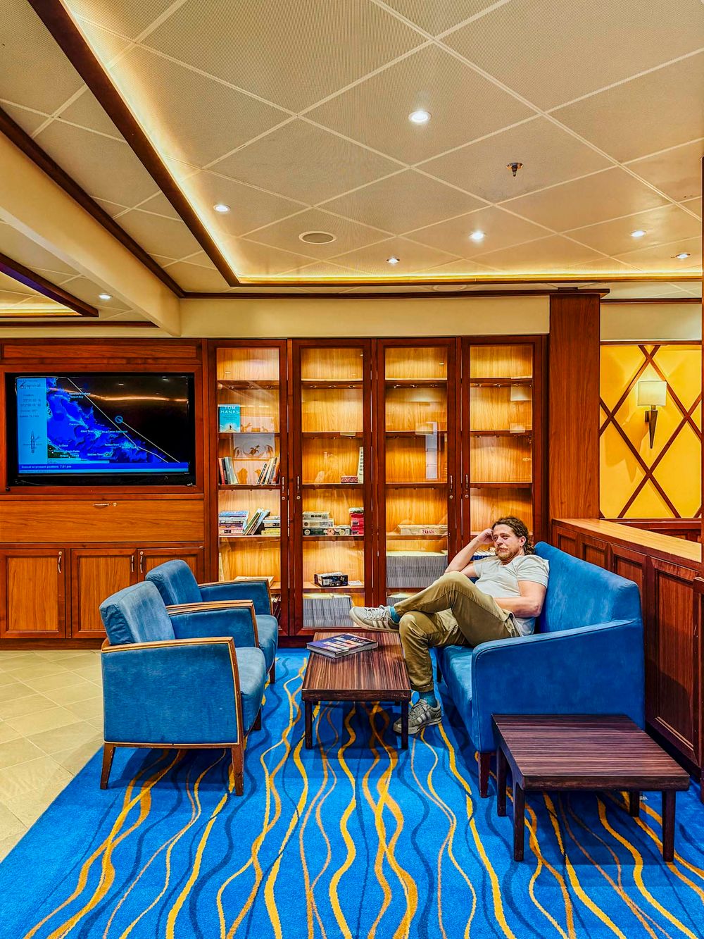 Man relaxing on blue velvet sofa in elegant cruise ship library lounge with wooden bookshelves and TV.