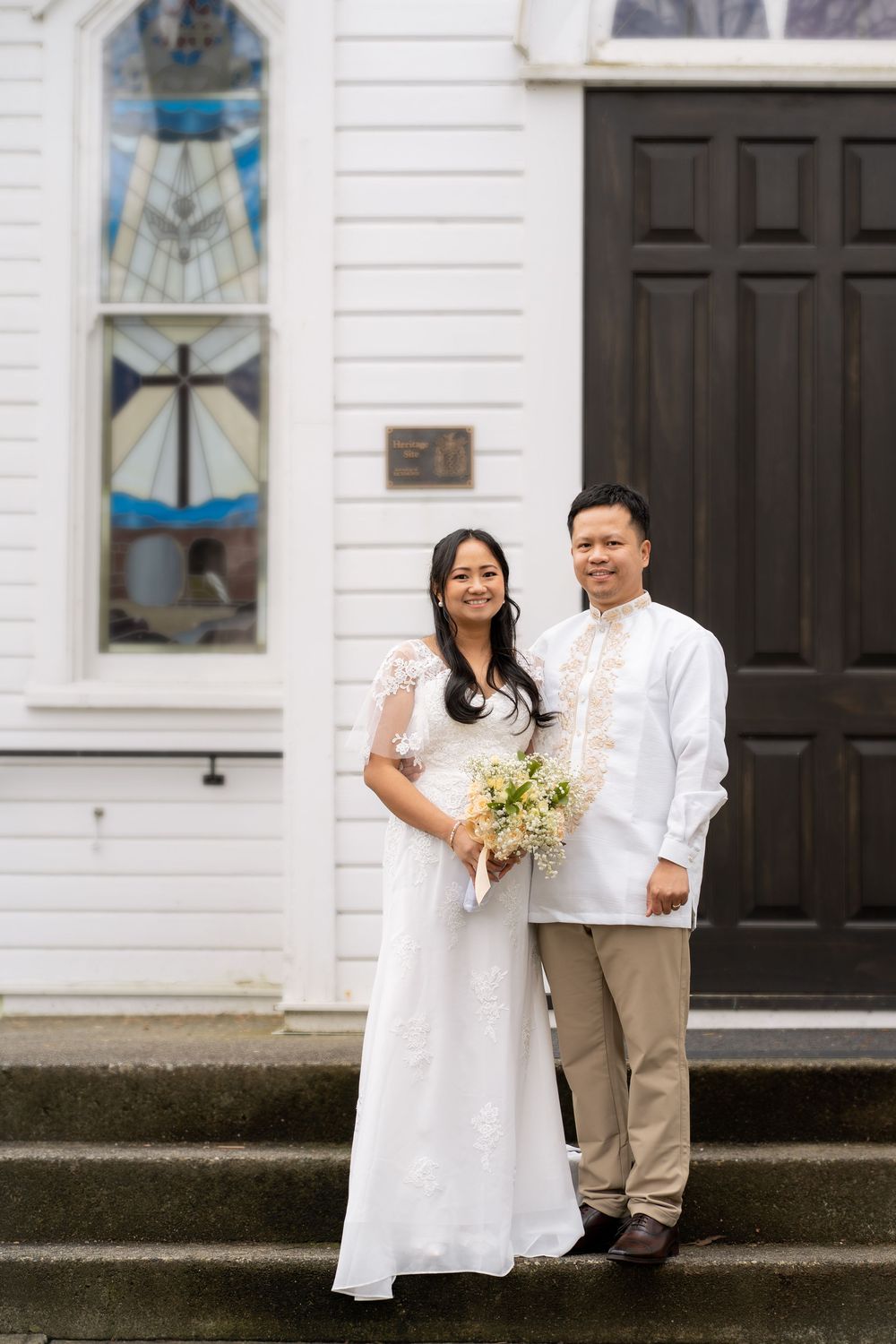 Asian couple in traditional wedding attire stands on church steps, bride holding white bouquet.