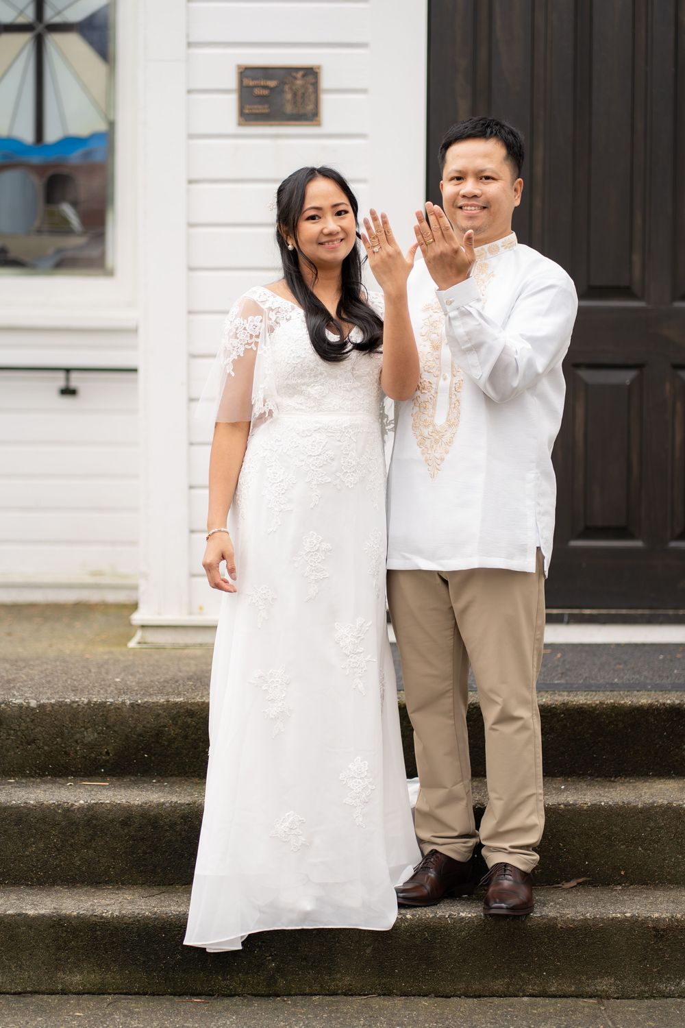 Asian couple in traditional wedding attire smiling and waving on church steps.