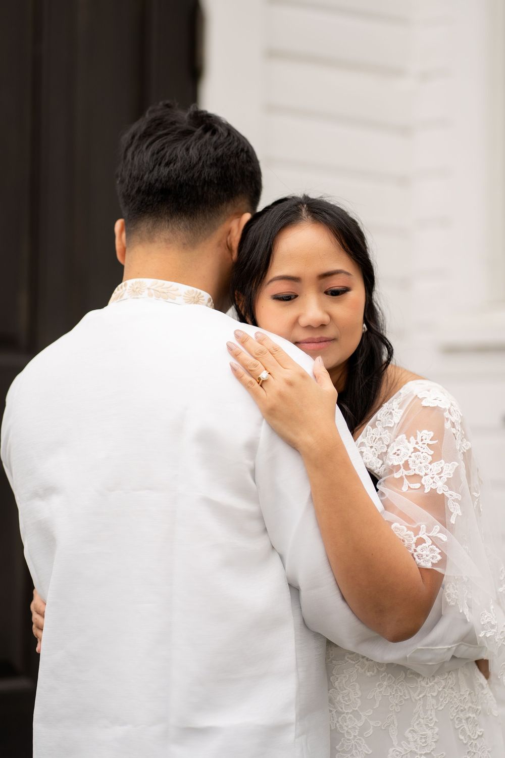 Asian bride and groom in white attire share an intimate embrace outside a white building on their wedding day.