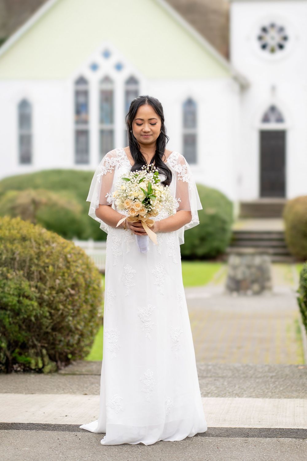 Bride in white lace gown holding bouquet outside a white church.
