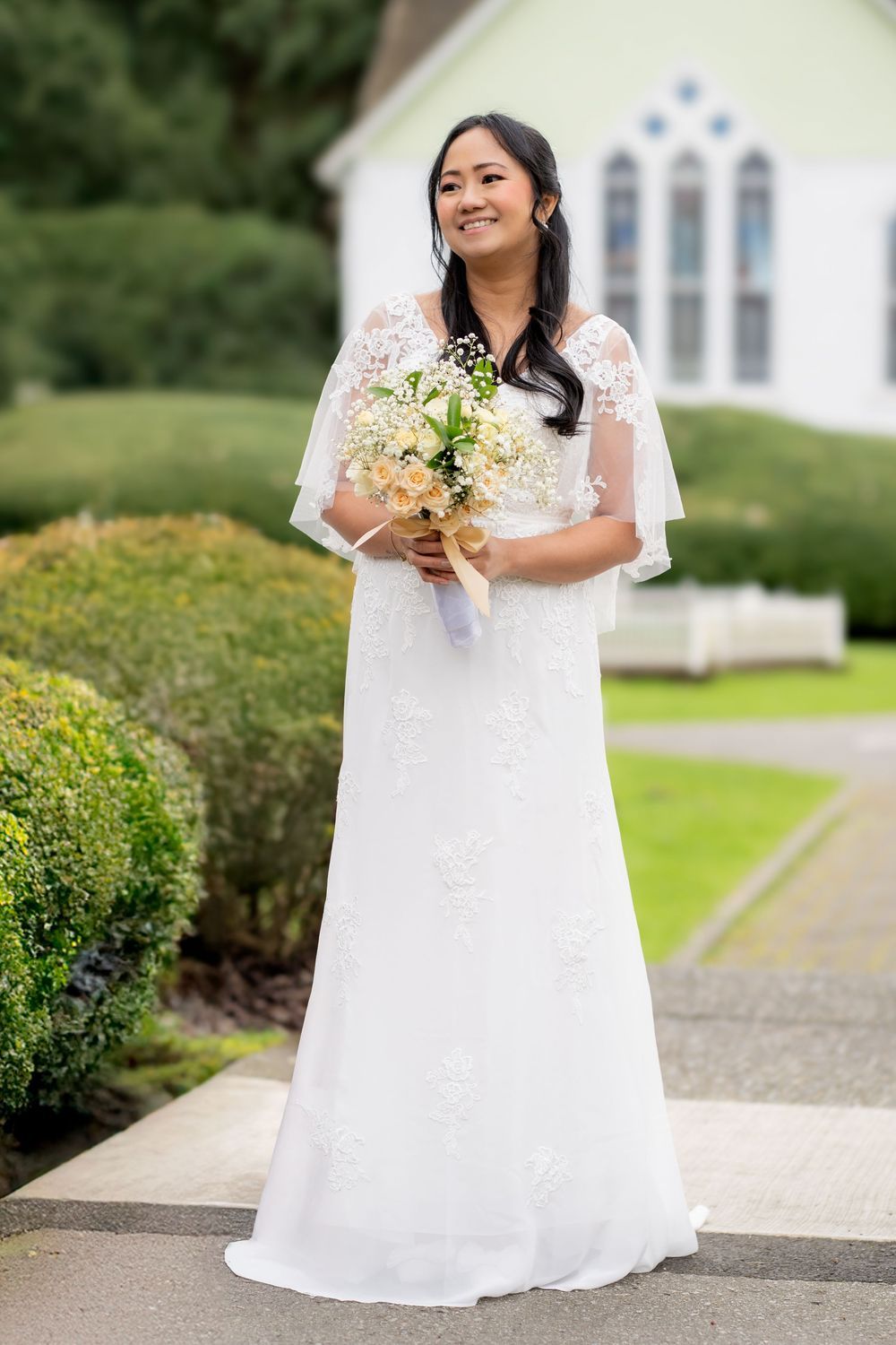 Smiling Asian bride in white lace gown holds bouquet outside a church surrounded by greenery.