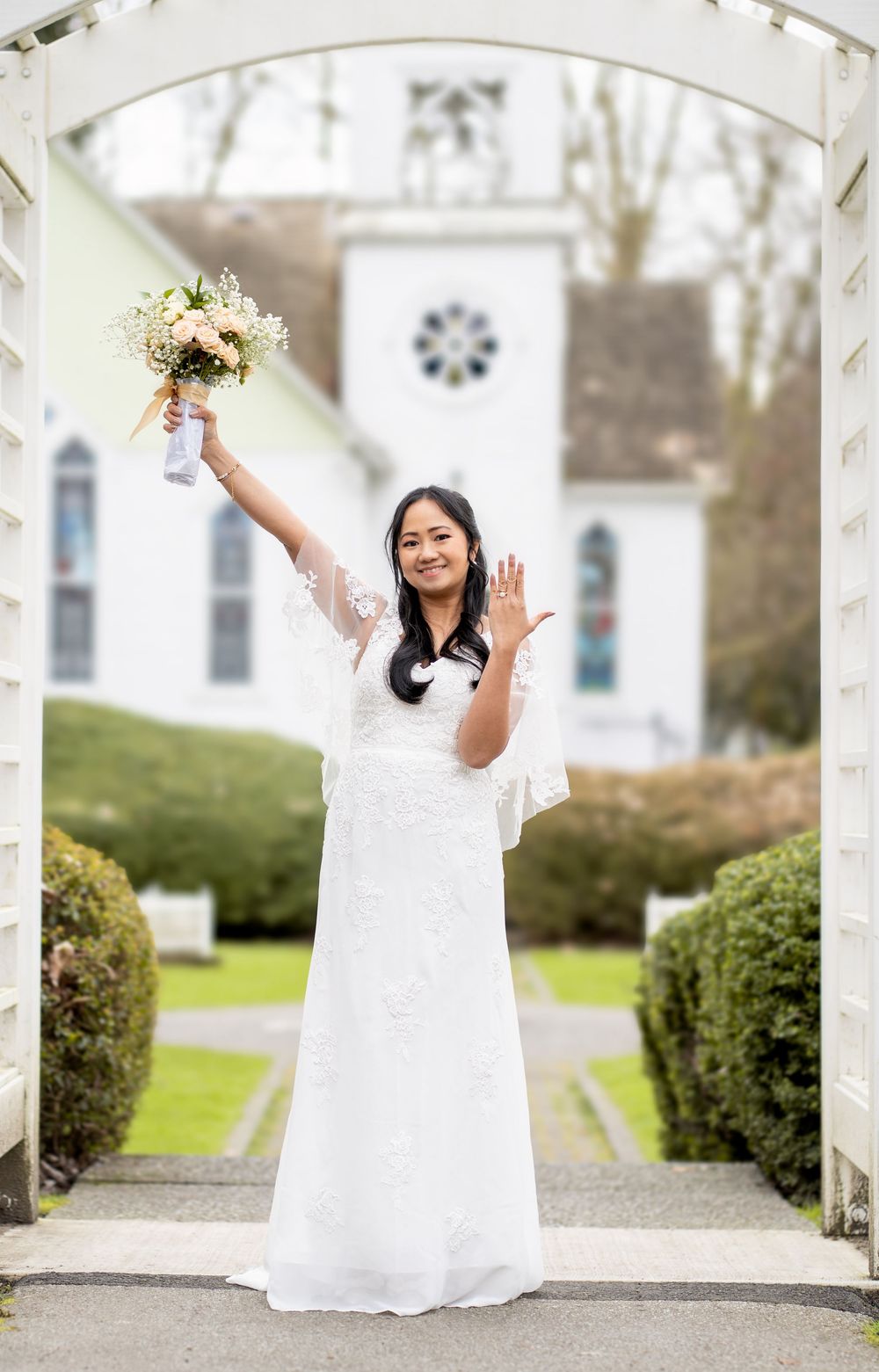 Smiling bride in white lace gown raises bouquet and waves outside a white church on her wedding day.