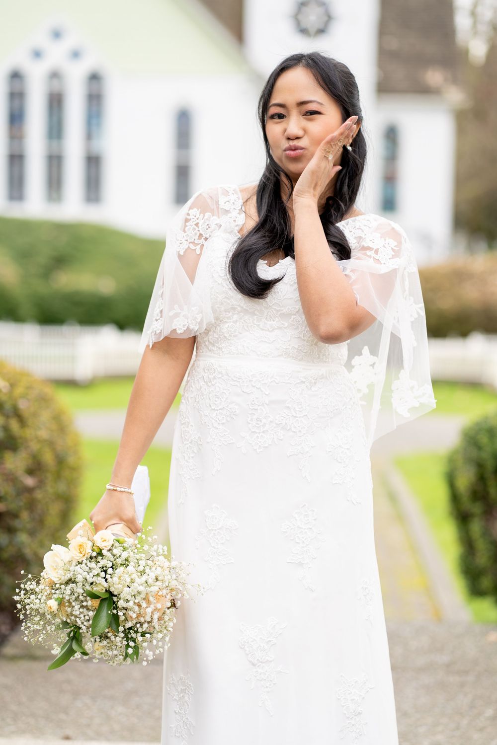 Asian bride in white lace gown blowing a kiss while holding a bouquet outside a church.