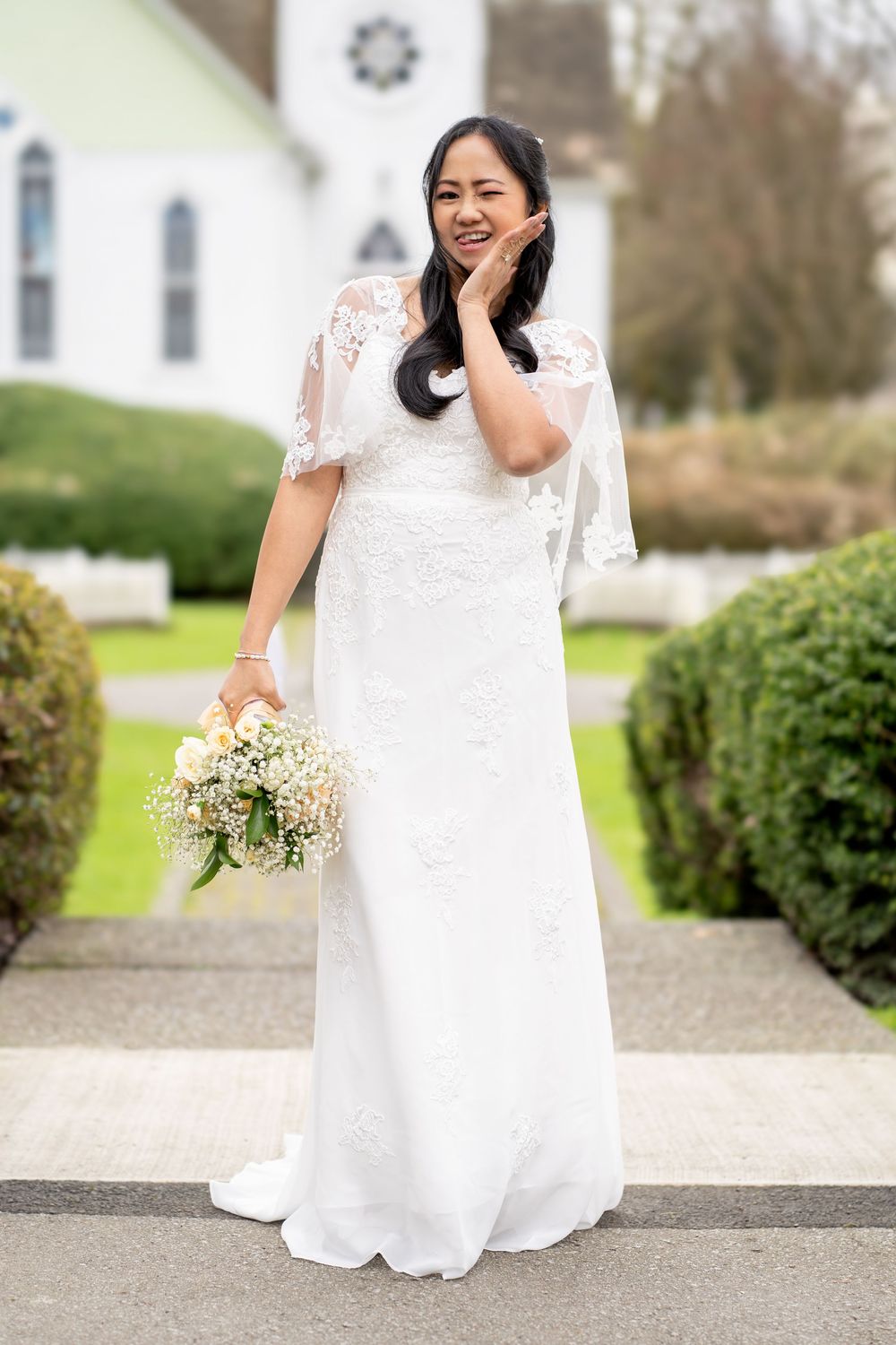 Smiling Asian bride in white lace flutter-sleeve gown holding bouquet, posing outdoors near a white church.