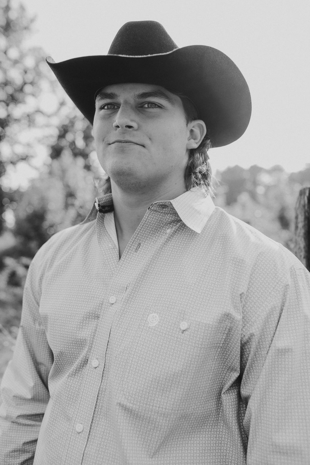 Black and white portrait of a young cowboy in a western hat and checkered button-up shirt looking off into the distance.