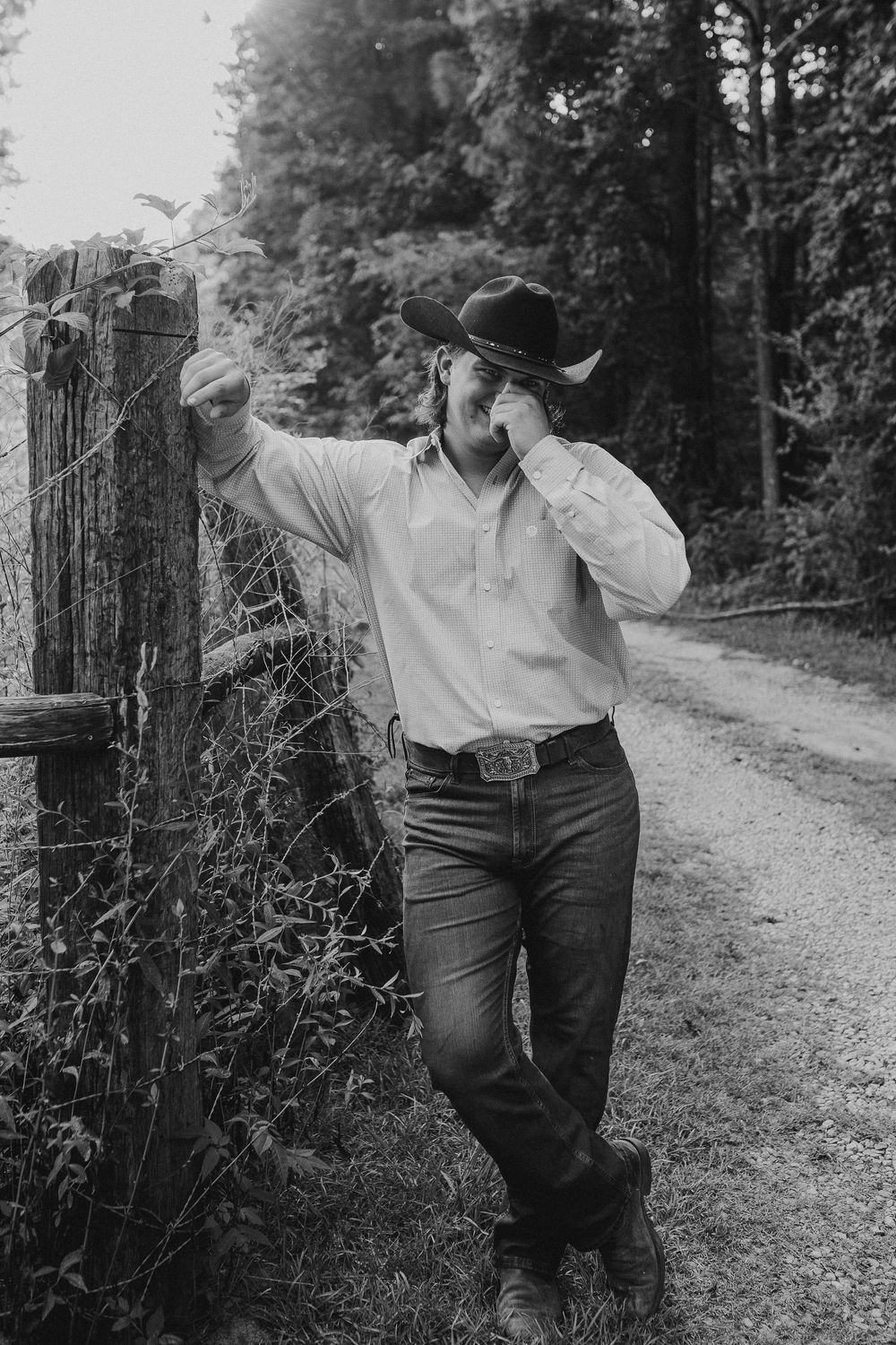 A couple embraces on a rustic dirt path lined with wooden fencing and lush greenery in a black and white photograph.