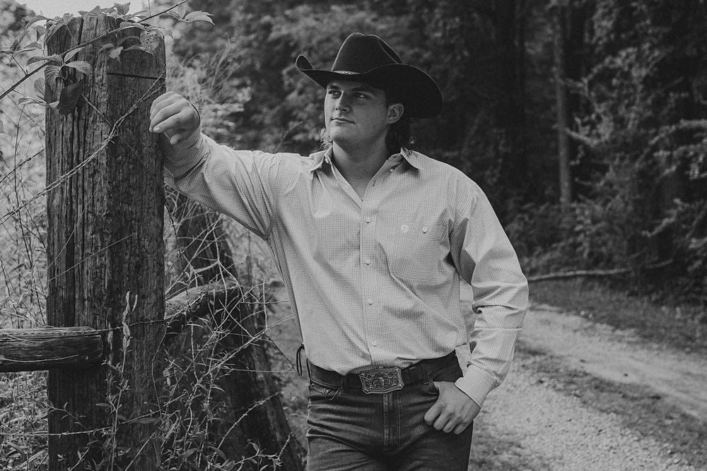 A person in a cowboy hat walks along a dirt path through a wooded area in black and white.
