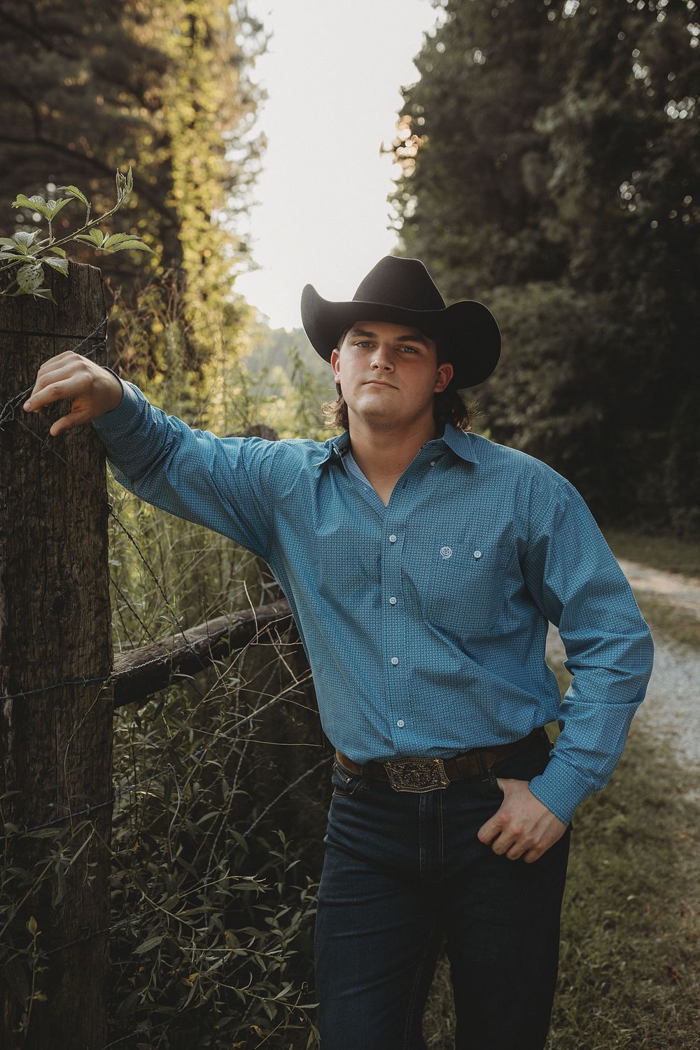 A person in a blue western shirt and black cowboy hat poses dramatically against a wooded outdoor setting.
