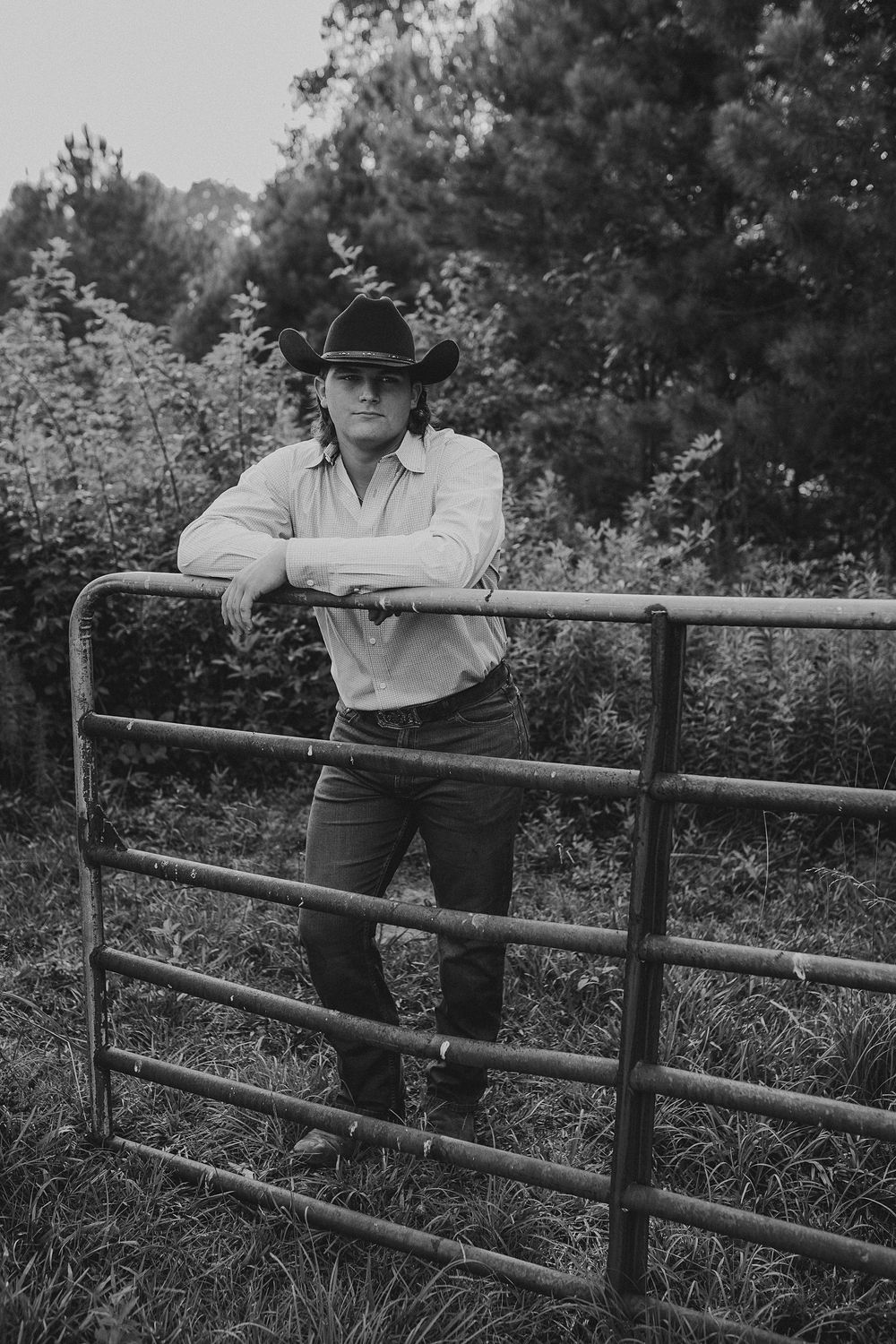 A person in a cowboy hat and western attire leans casually against a wooden fence in a rural outdoor setting.