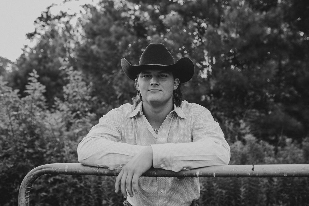 Black and white portrait of someone in a cowboy hat leaning on a fence post in an outdoor setting.