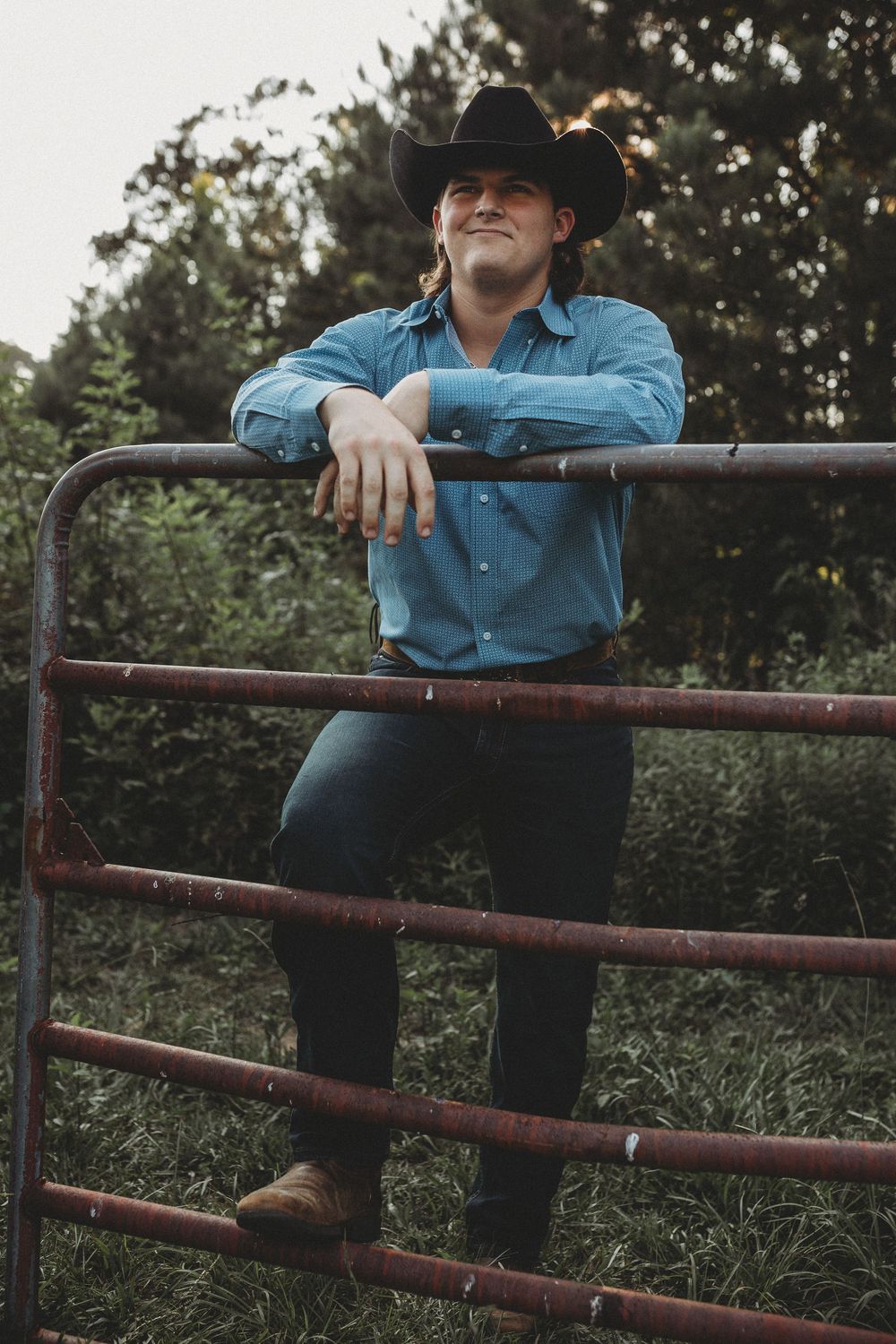 A cowboy in a blue denim shirt leans on a rustic metal gate in a rural outdoor setting.