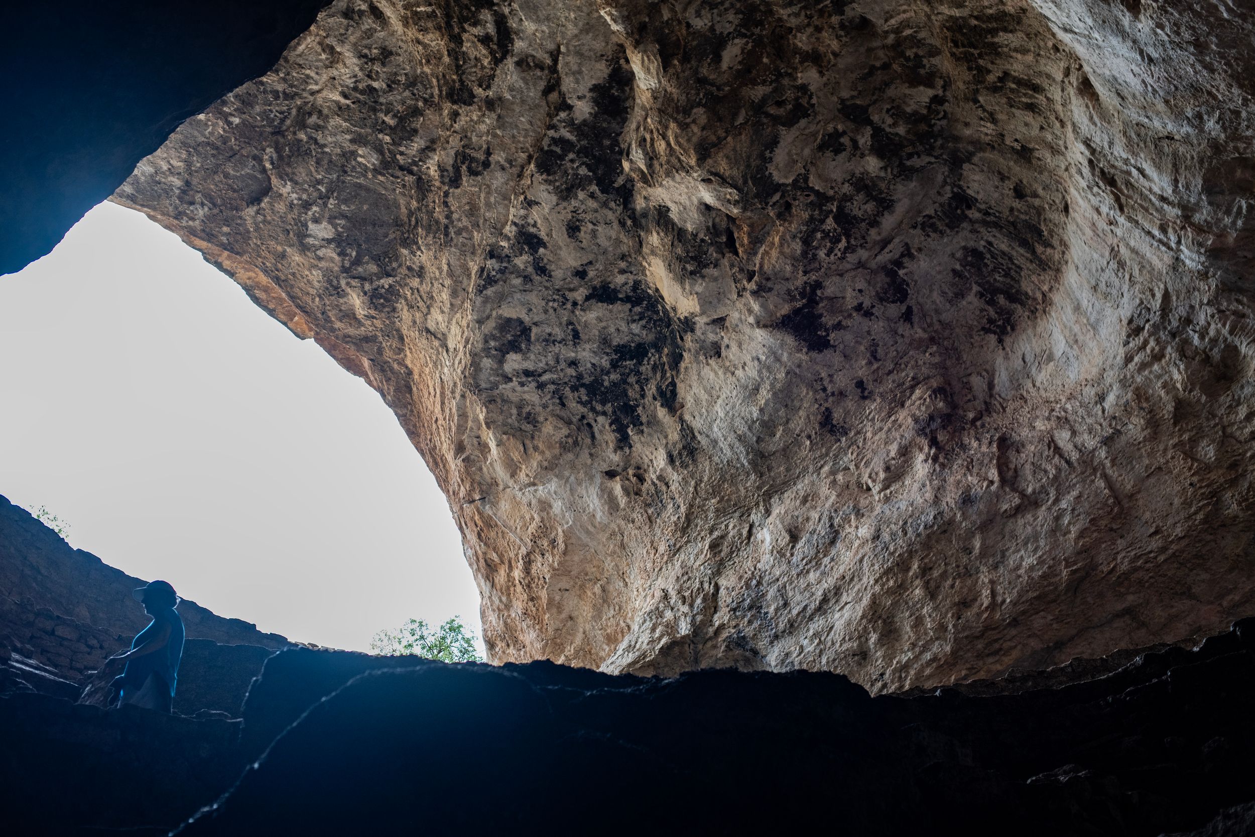 View from inside a dark cave looking up at a large rocky opening with bright natural light illuminating the stone ceiling.