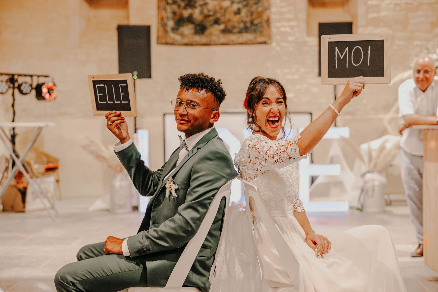 A joyful newlywed couple sits back-to-back holding chalkboard signs reading ELLE and MOI at their elegant wedding reception.