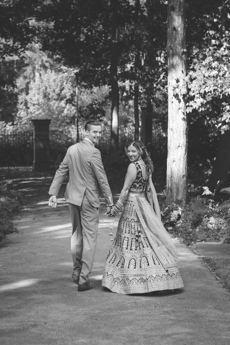 Black and white photo of a couple in wedding attire holding hands,walking along a tree-lined path with the bride wearing traditional dress