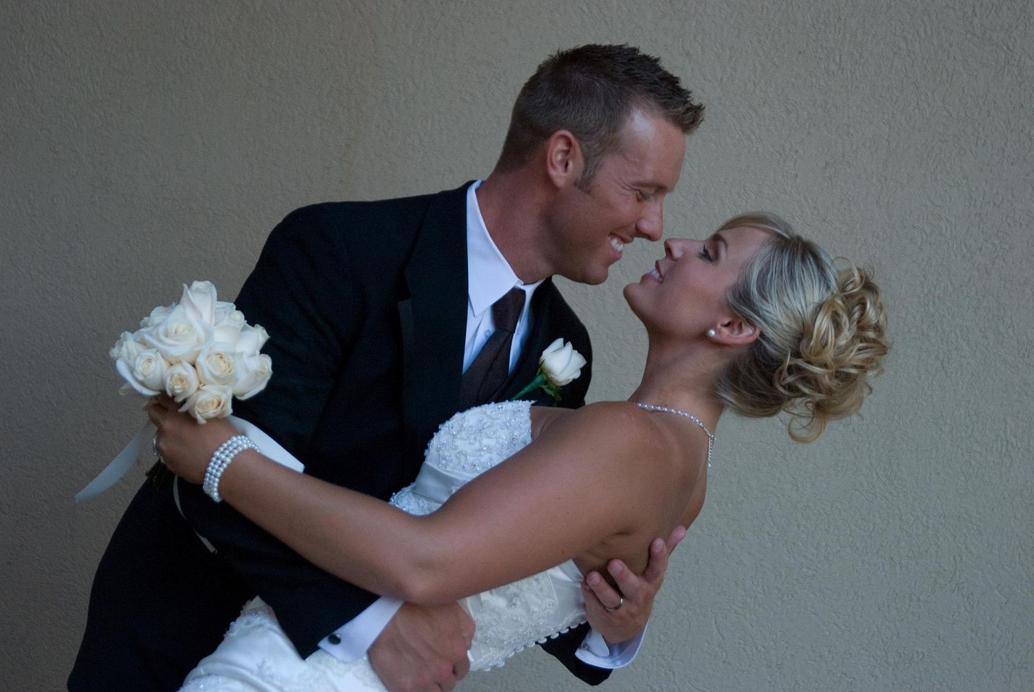 Groom in black suit dips bride in white strapless gown holding white bouquet during romantic wedding photo.