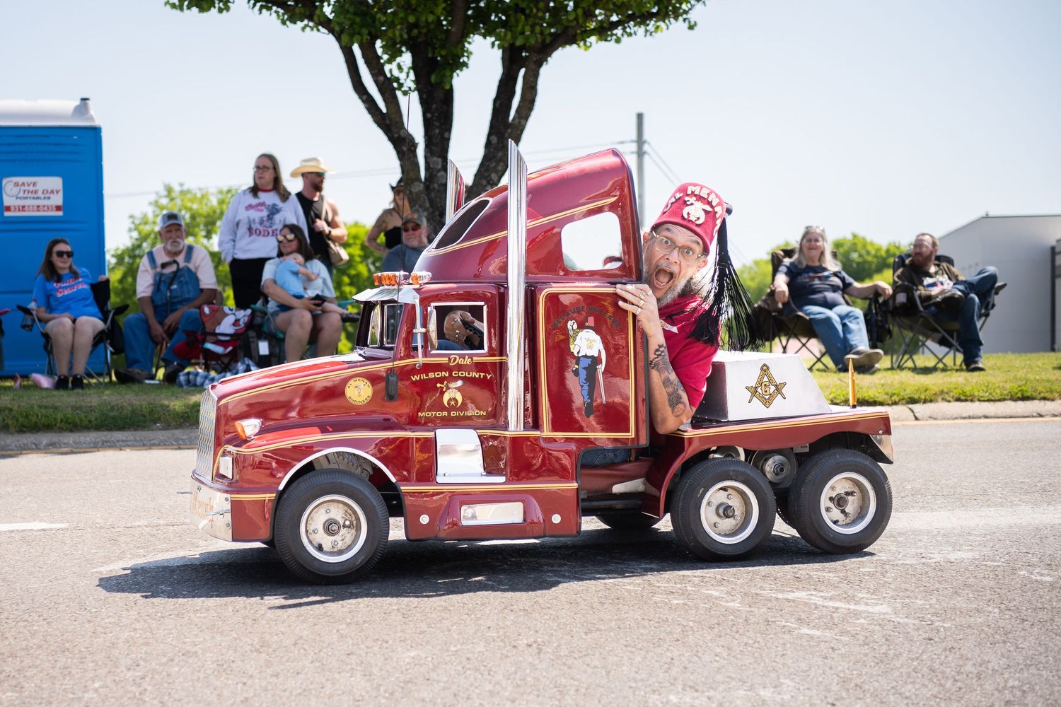 A miniature red semi-truck driven by a Shriner in a parade, with spectators watching from the sidewalk.