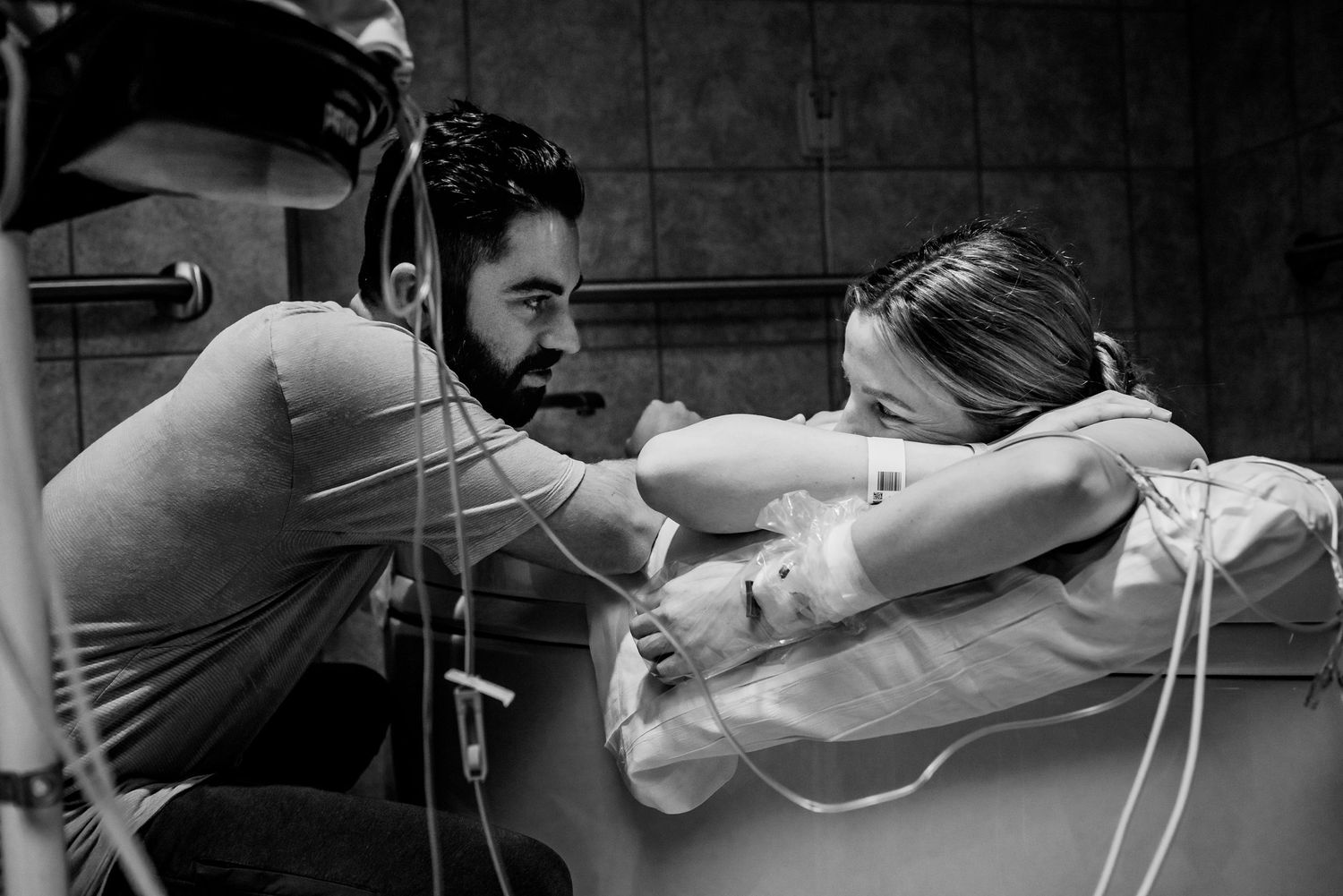 Black and white photo of a man supporting a woman during labor in a bathtub, connected to medical tubes.