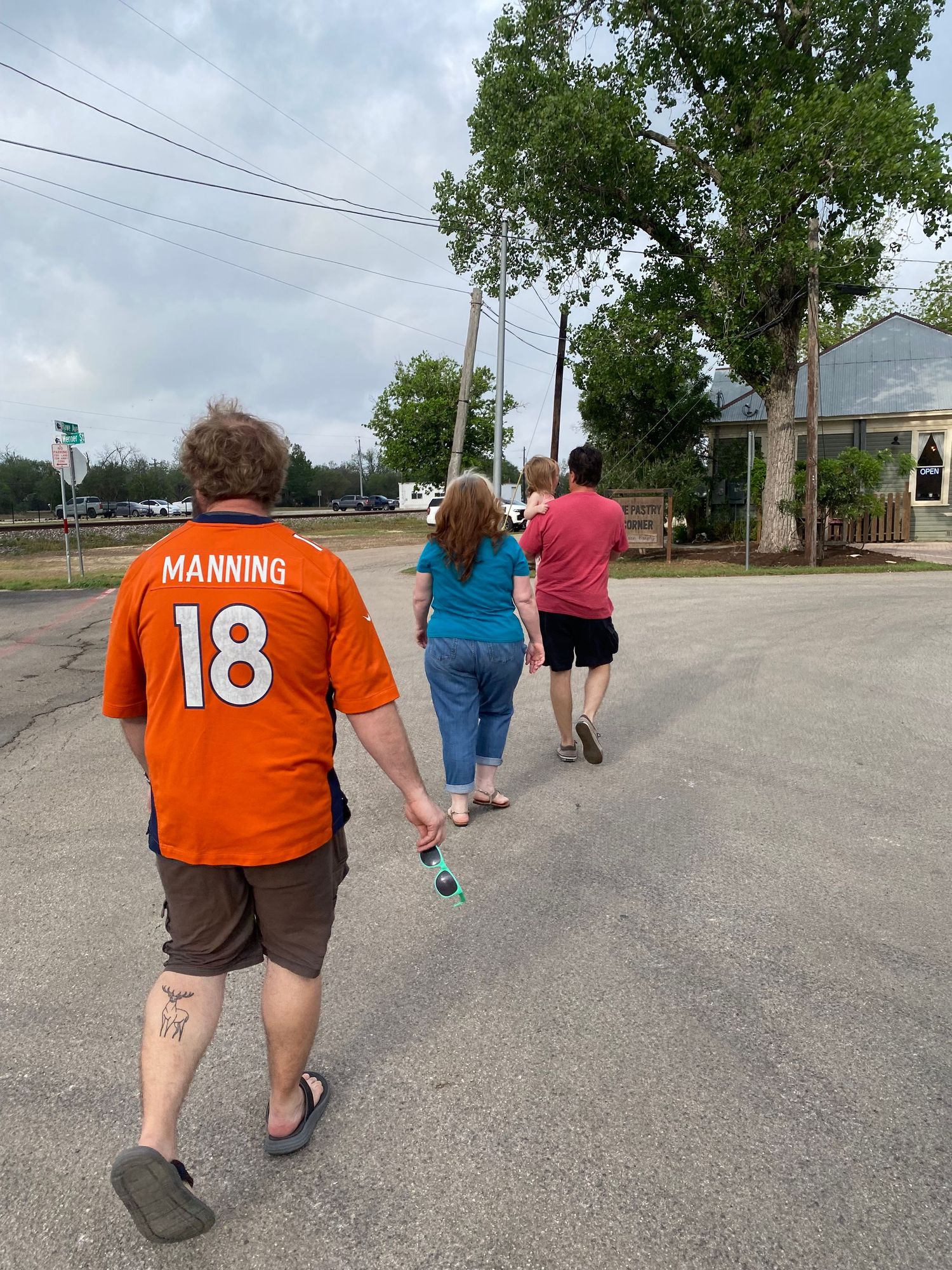 Three people walking away on a street, one wearing an orange Peyton Manning #18 Broncos jersey, on a cloudy day.