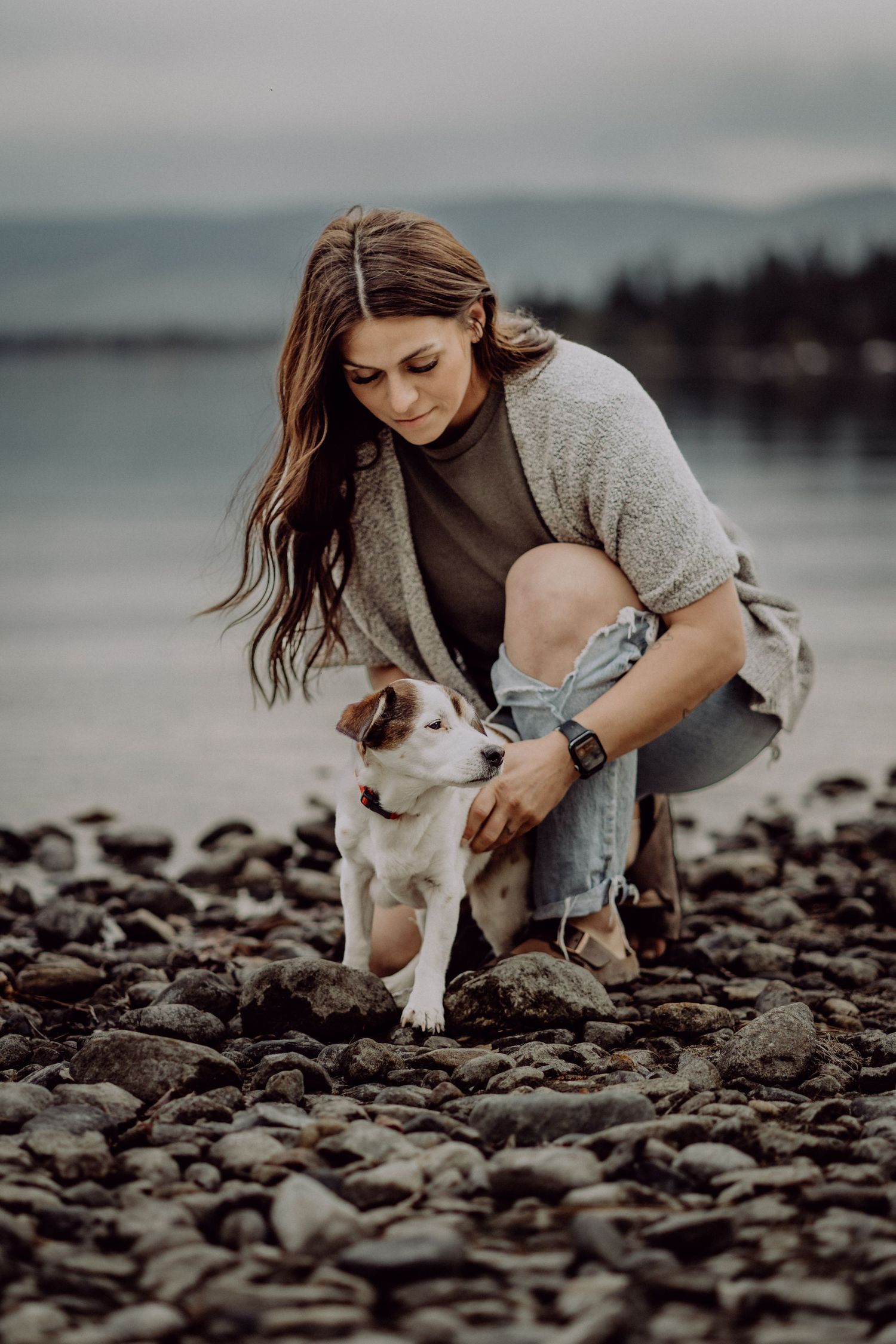 Woman crouching on rocky beach petting a Jack Russell Terrier dog near calm water on overcast day.
