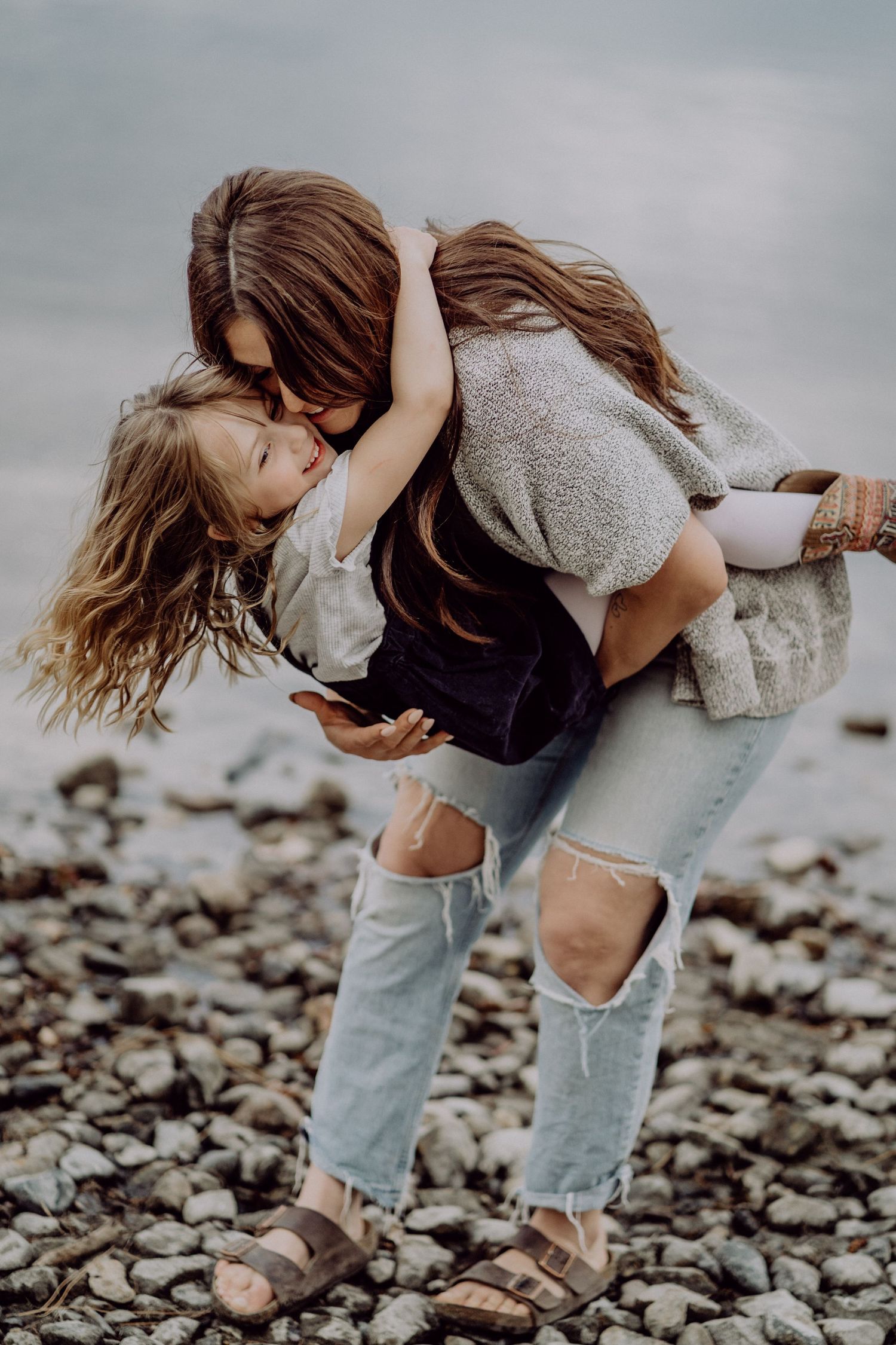 A mother playfully lifts her young daughter on a rocky beach, both laughing and embracing in casual bohemian outfits.