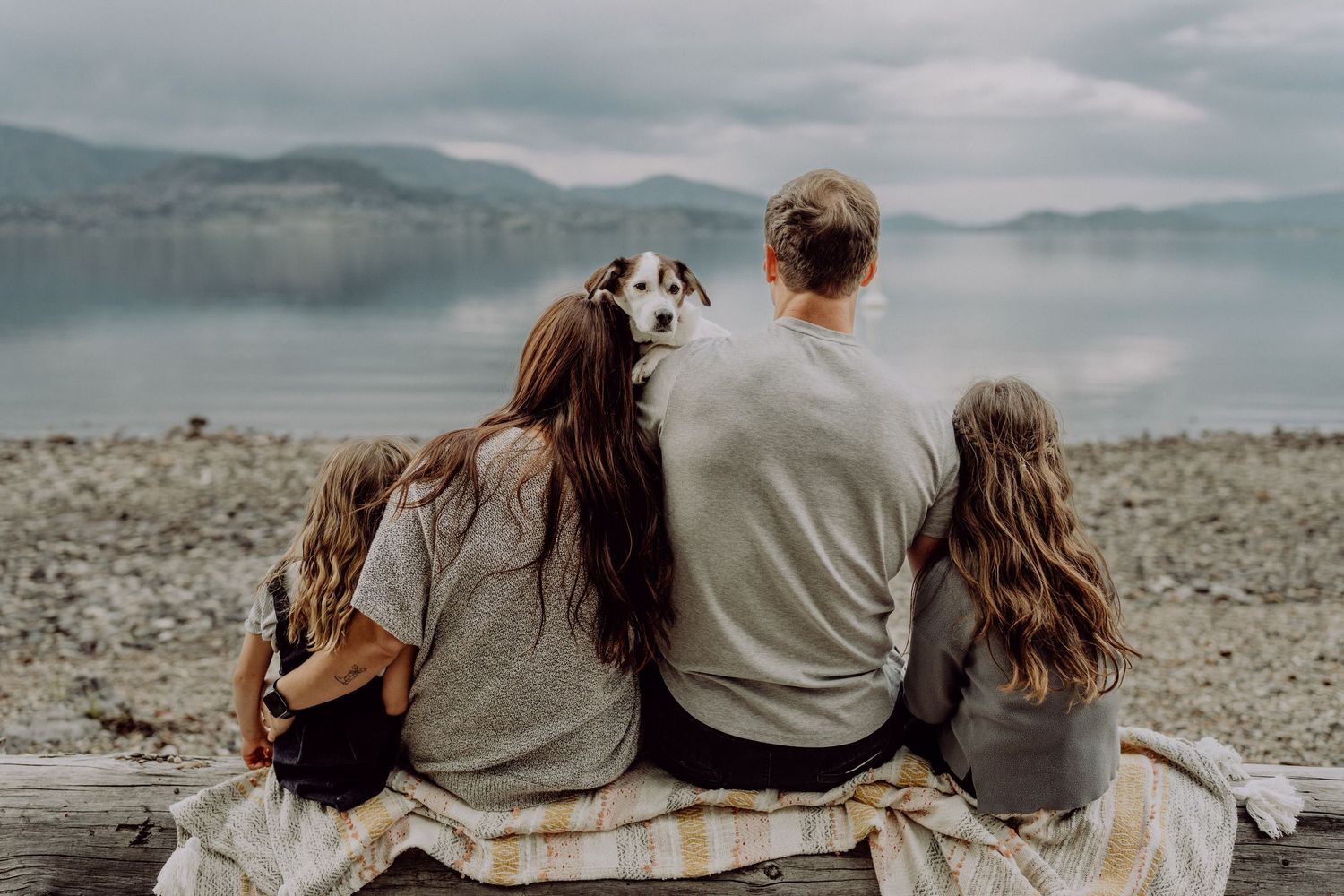 Family with two girls and a dog sitting on a bench by a scenic lake, viewed from behind on a cloudy day.