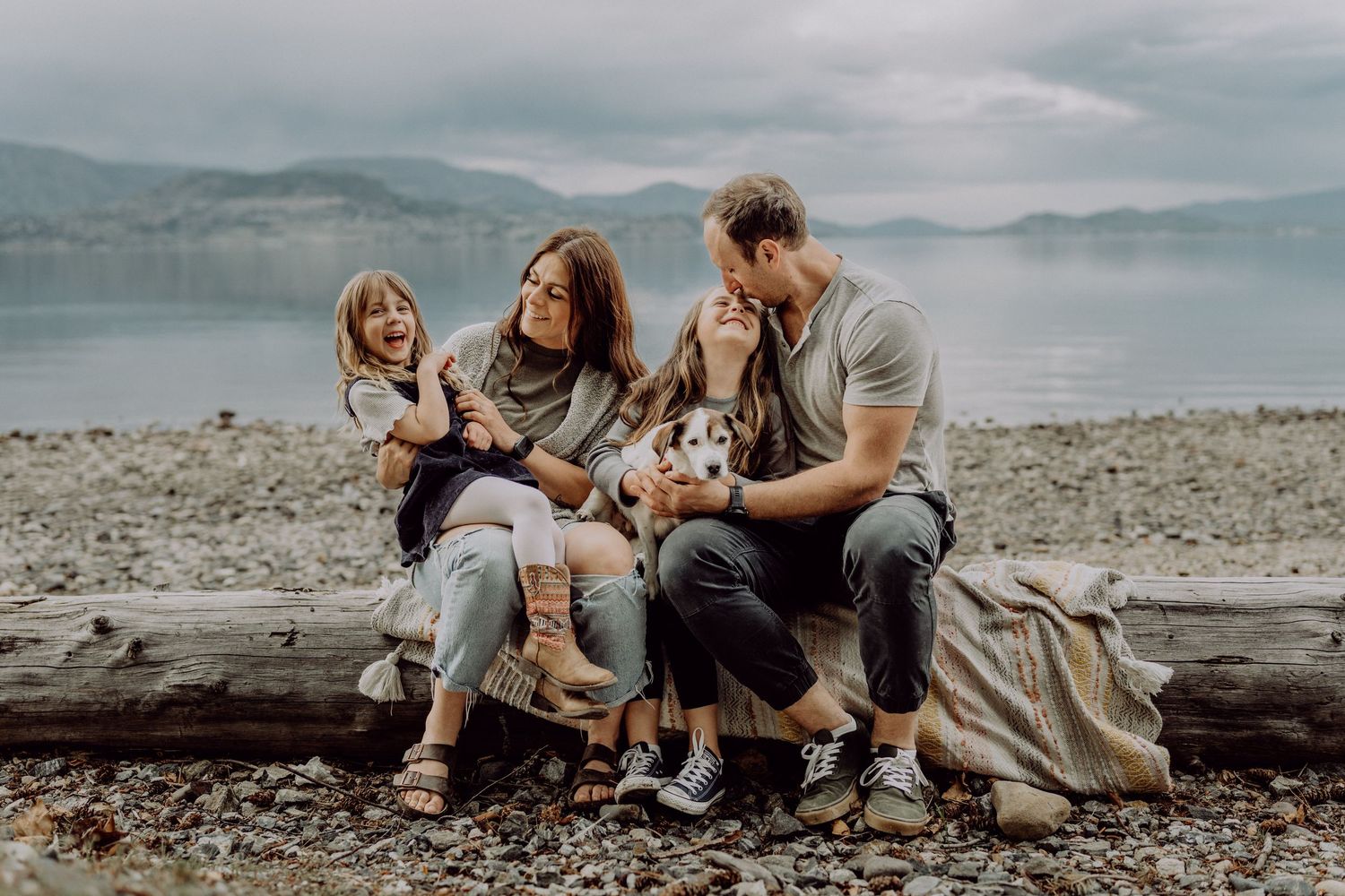 Family of four with a dog sitting on a driftwood log on a rocky beach with mountains and lake in background.