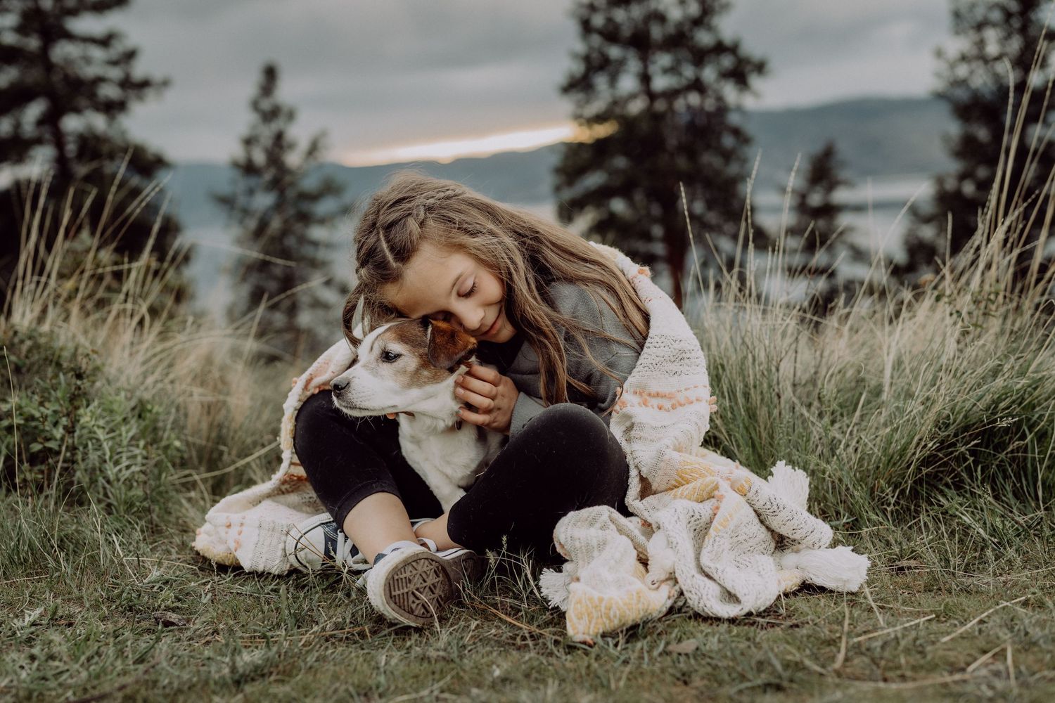 Young girl sitting on grass hugging a white dog with pine trees and cloudy sky in the background.