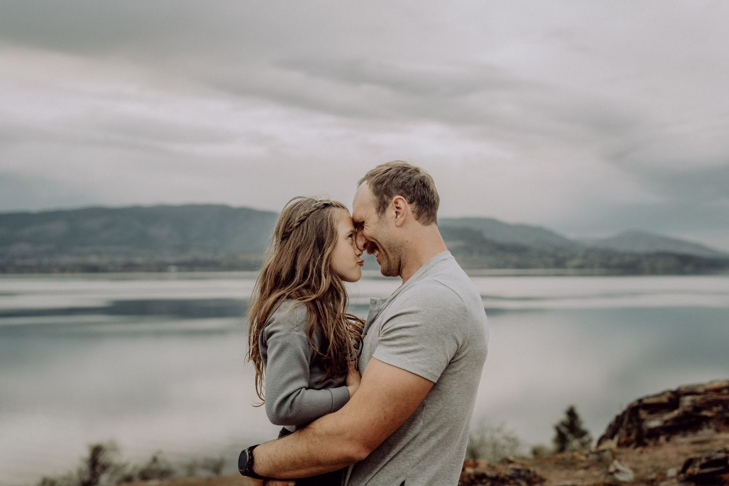 A couple embracing and touching foreheads romantically with a scenic lake and cloudy sky behind them.