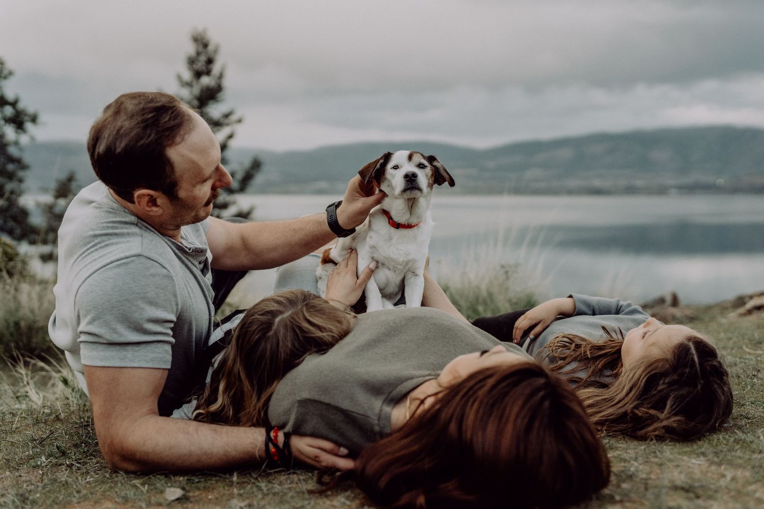 Family relaxing outdoors by a lake with their beagle dog, enjoying a peaceful moment together on the grass.