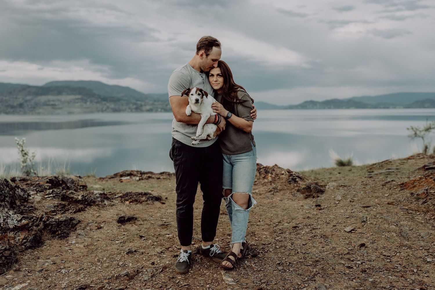 Couple holding a small dog on a rocky overlook with a serene lake and misty mountains in the background.