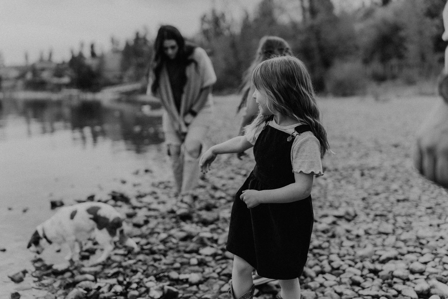 Black and white photo of a young girl in a dress playing on a rocky shoreline while family members explore nearby.