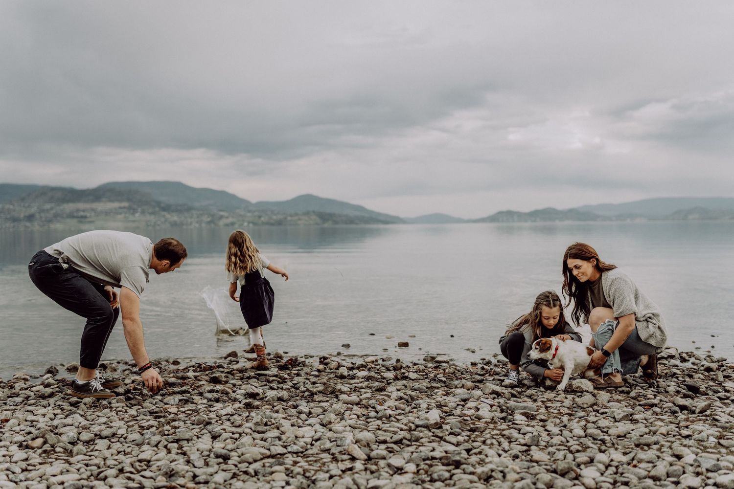 Family of four playing together on a rocky beach shore with mountains and cloudy sky in the background.