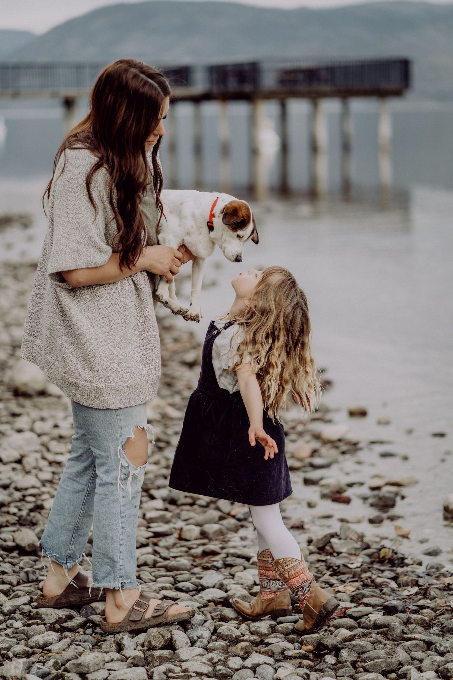 A mother holds a small dog while her young daughter leans in for a kiss on a rocky beach near a pier.