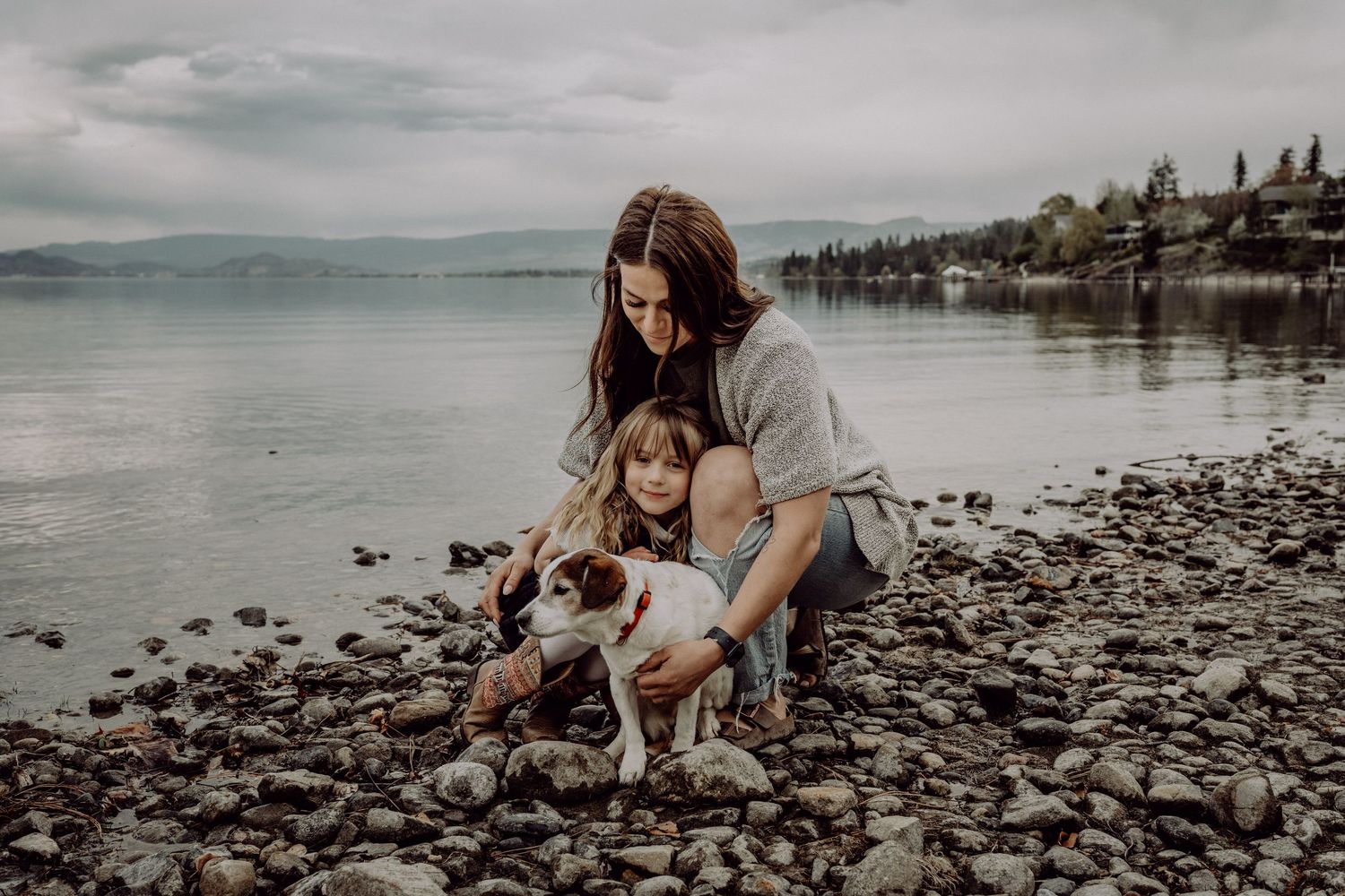 A mother and young daughter kneel on a rocky beach petting a white and brown dog by a serene lake.