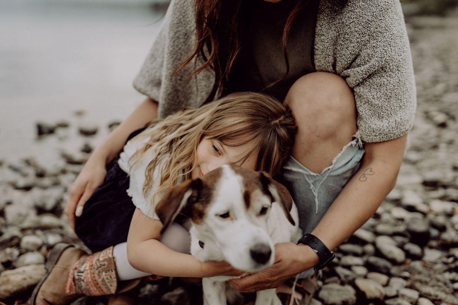 A young girl and woman cuddle a brown and white Husky dog on a rocky beach shore.