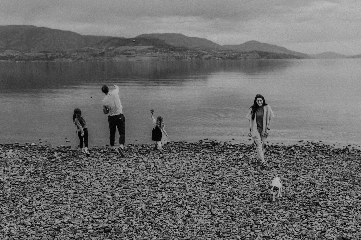 Black and white photo of a family playing on a rocky beach beside calm water with misty mountains in the background.