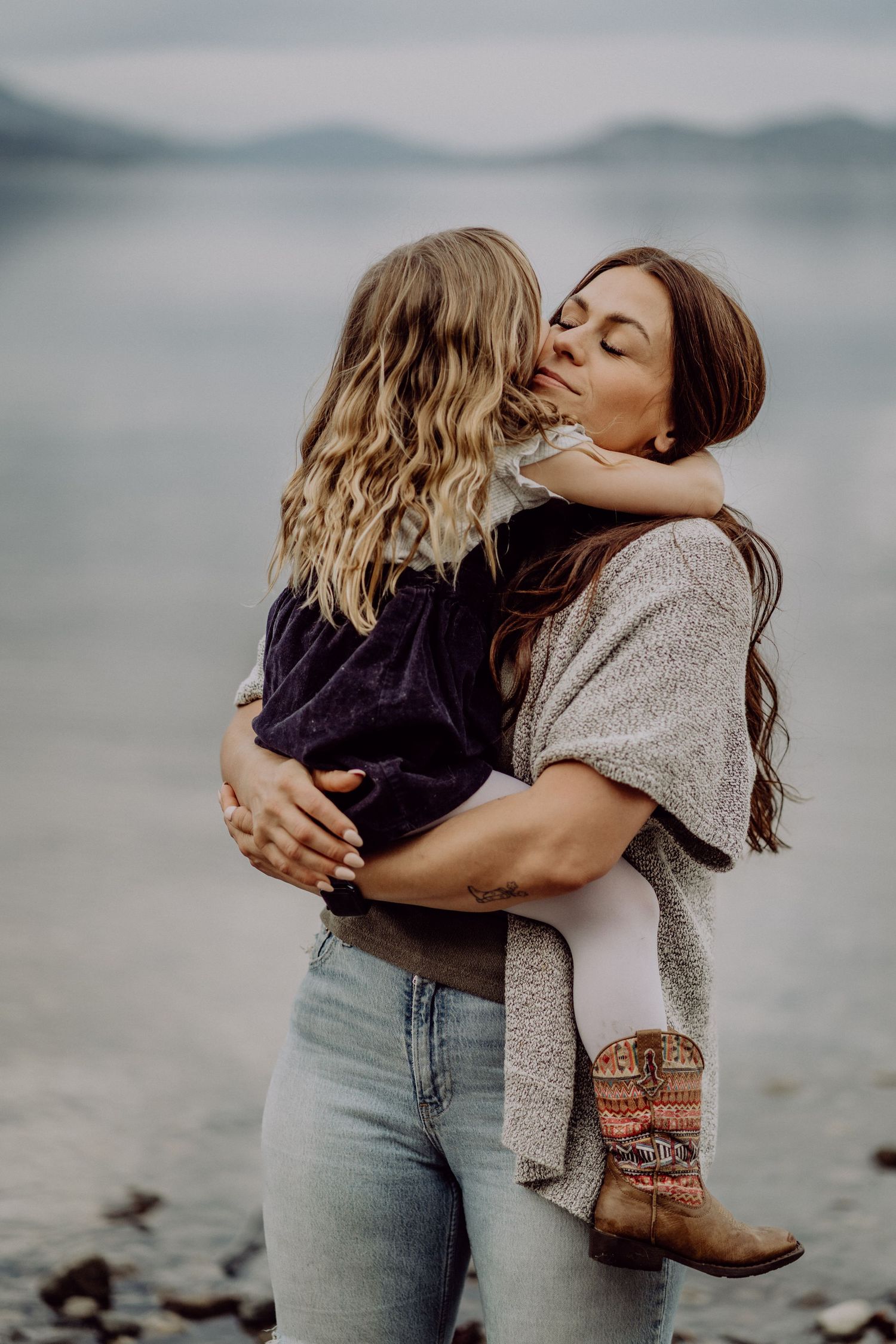 A mother tenderly kisses her young daughter while holding her on a misty lakeside beach.