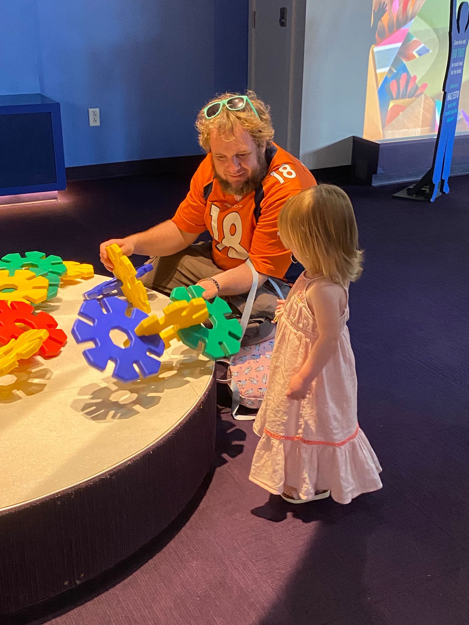 A father in an orange Broncos jersey plays with colorful gear toys alongside his toddler daughter at a children's museum.
