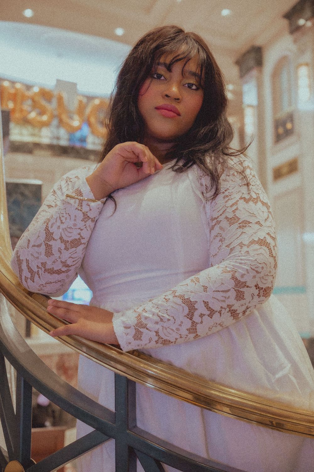 A young woman in a white lace dress poses gracefully on a golden staircase railing indoors.