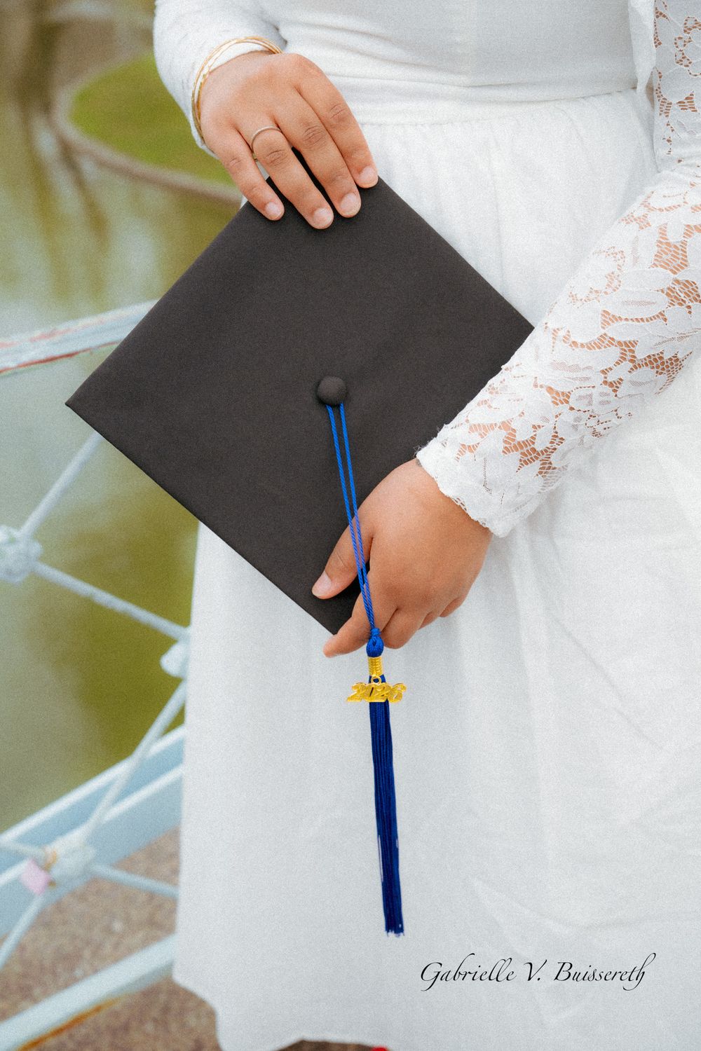 A graduate holds a black mortarboard cap with blue tassel while wearing a white lace dress.