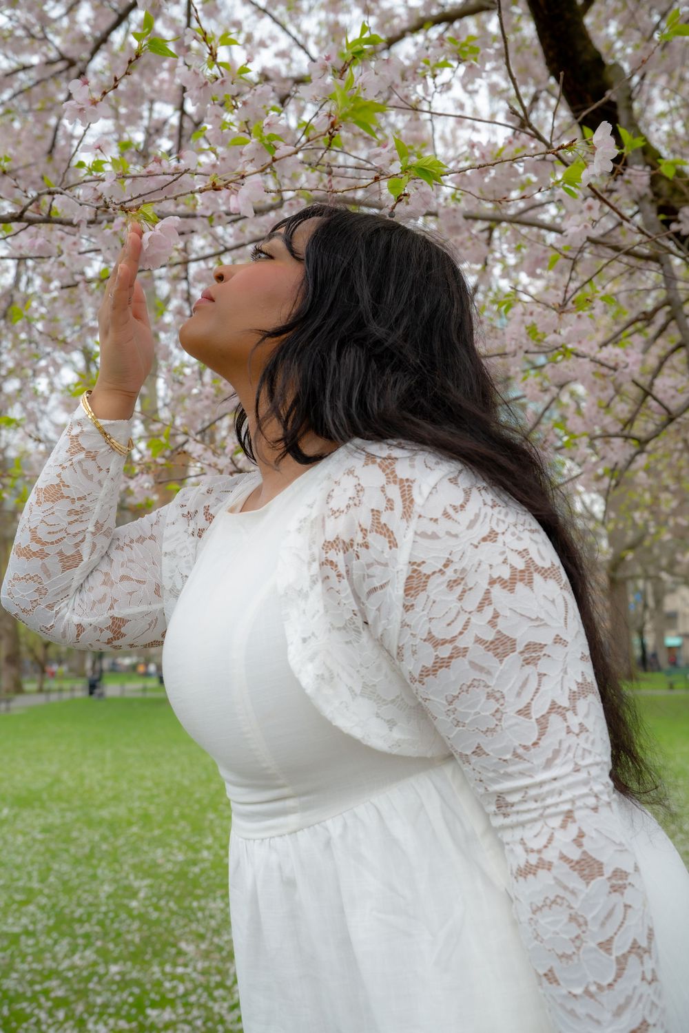Young woman in white lace dress reaching up to touch pink cherry blossom tree in park.