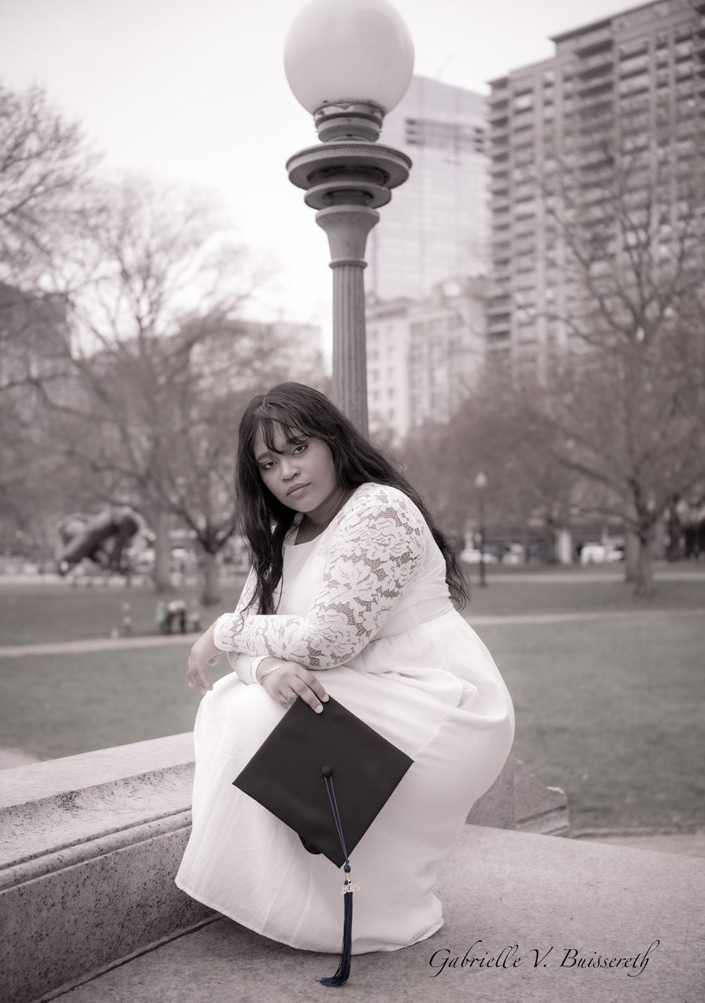 Graduate in white lace dress holding black mortarboard cap seated on stone ledge in park.