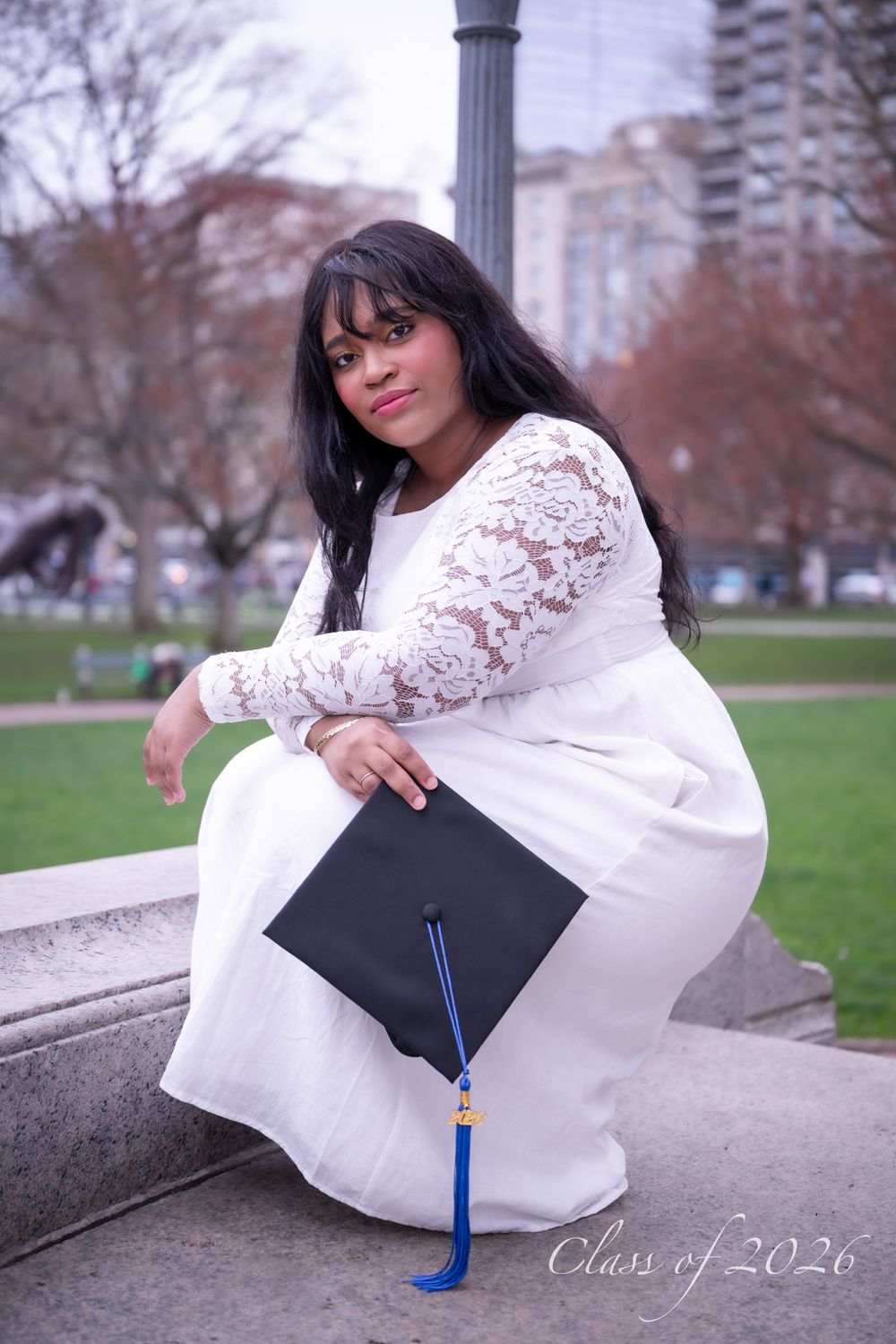 Young graduate in white lace dress holding black cap with blue tassel seated outdoors in park.