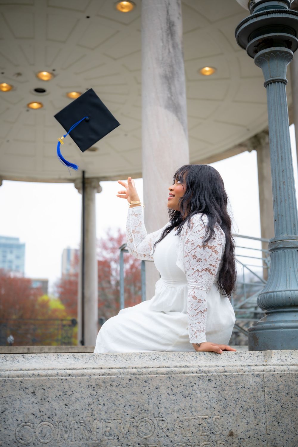 Graduate in white lace dress tossing mortarboard cap in air at outdoor pavilion celebration.