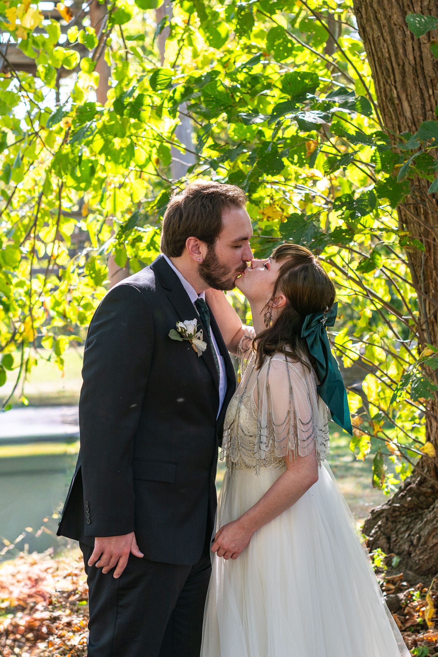 A bride and groom share a romantic kiss under lush green tree branches during their outdoor wedding ceremony.