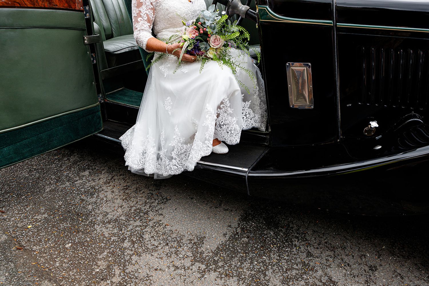 Bride in lace wedding dress holding bouquet, seated on vintage black car step on rainy day.